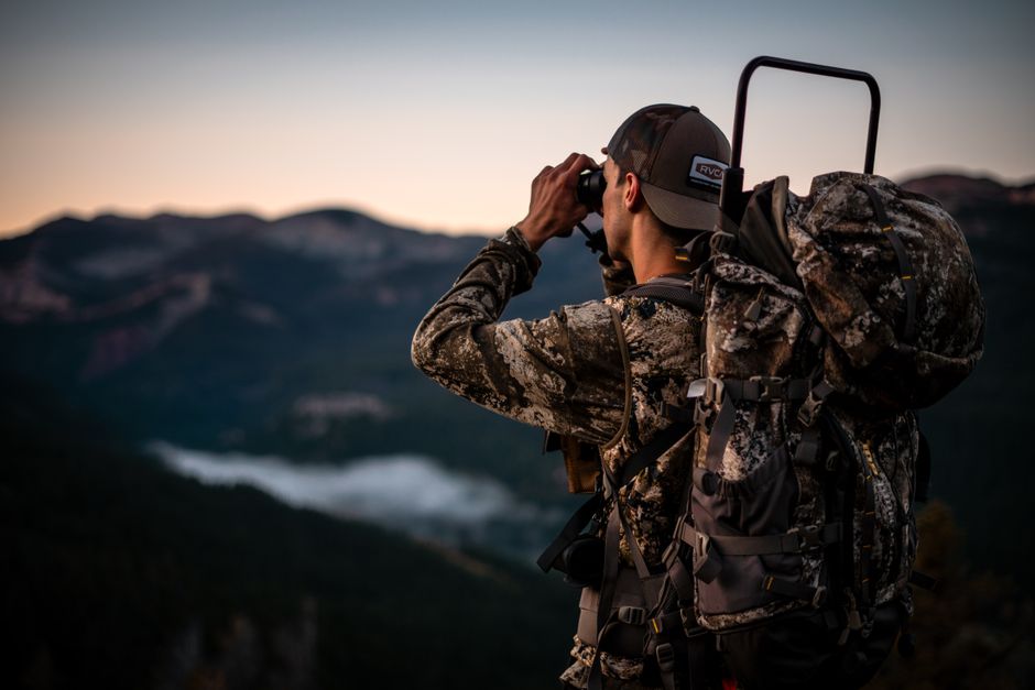 Camouflaged hiker with backpack using binoculars in scenic mountain landscape at dusk.